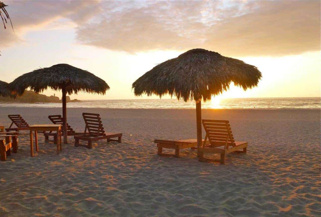 beach at sunset in golden light with palmtree umbrellas and a few beach beds made of wood