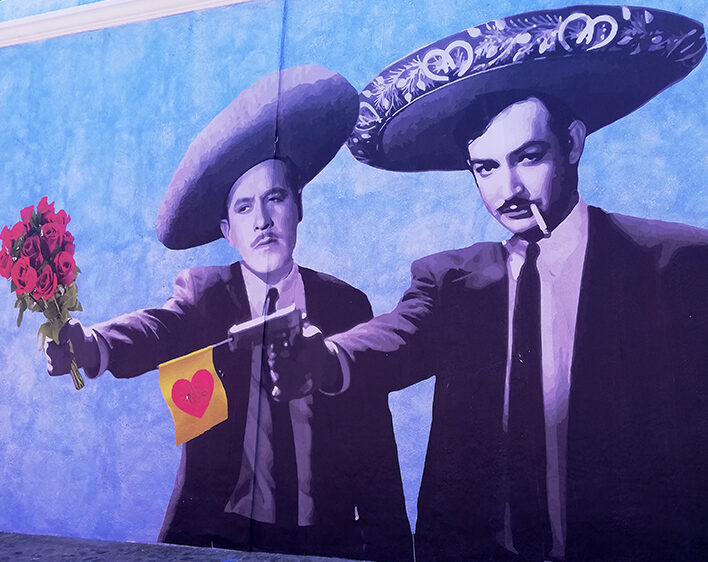 Street with two men wearing Mexican sombreros and holding a flower bouquet as well as a pistol with a heart flag