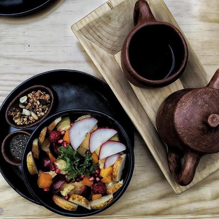 Breakfast granola with fruits in a black bowl next to a tea pot and tea cup on a wood plate