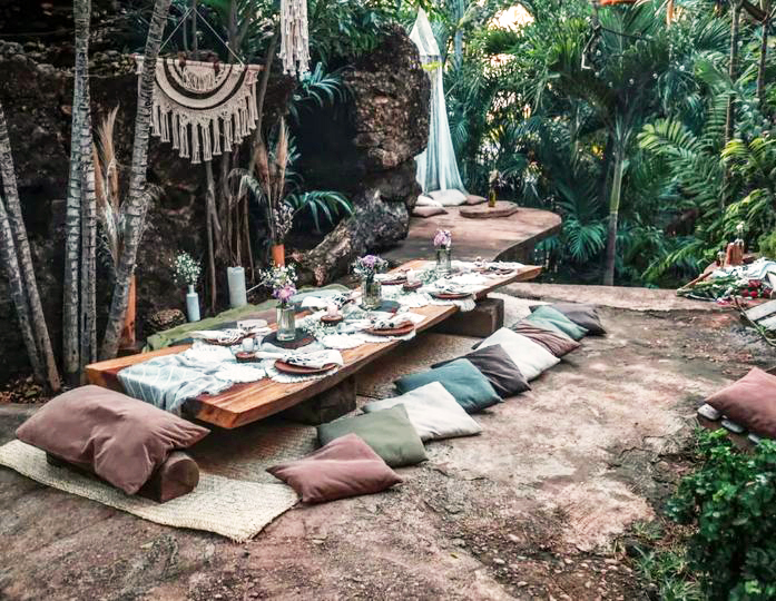 wooden table low on the floor surrounded by nature on an earthen ground, a few suede pillows around the table, and the table set with flowers, decoration and ceramic plates