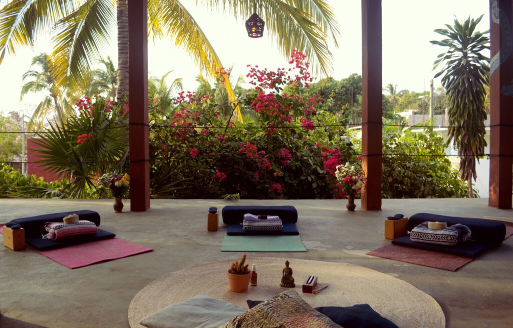 Open air yoga studio surrounded by plam trees, with a ypga bolster and some yoga mats laid out on the floor