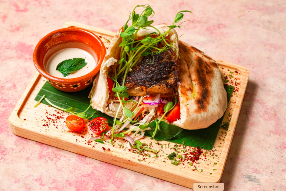 vegan pitta bread filled with tomatoes, lettuce, and falafel, on a banana leaf on a wooden plate, with a small terracotta cup of sauce next to it