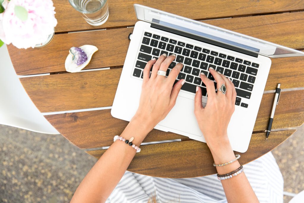 laptop on a wooden table, women with hands on the laptop wearing crystal bracelets, a pen next to the laptop, an amethyst crystal on the other side