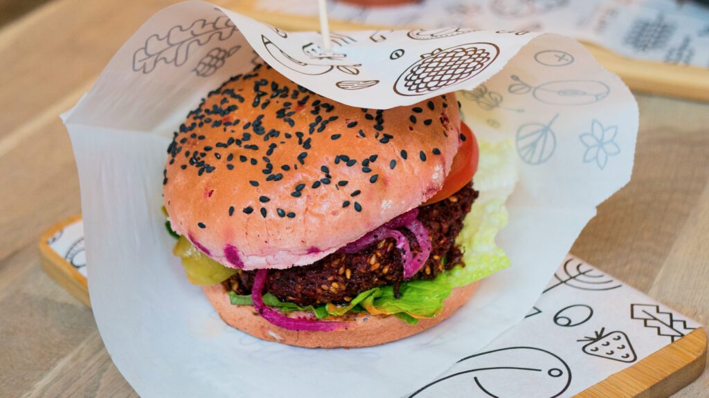 vegan burger on a wooden tablet and a wooden stick holding it together, wrapped with paper that has vegetables printed on it
