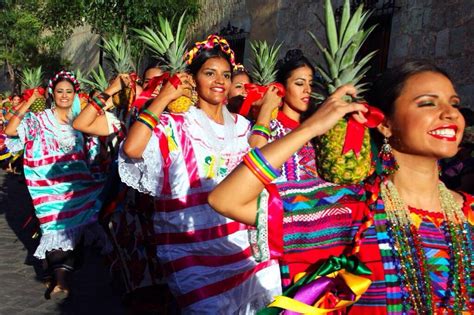 Women in Oaxaca, Mexico, dressed in colorful dresses, holding a pineapple on their shoulder as part of a procession on the streets