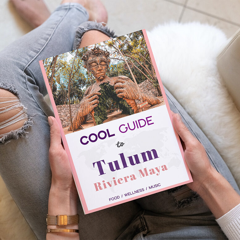 woman in ripped jeans sitting in creme-colored armchair with a guide book that reads "tulum" in her hands